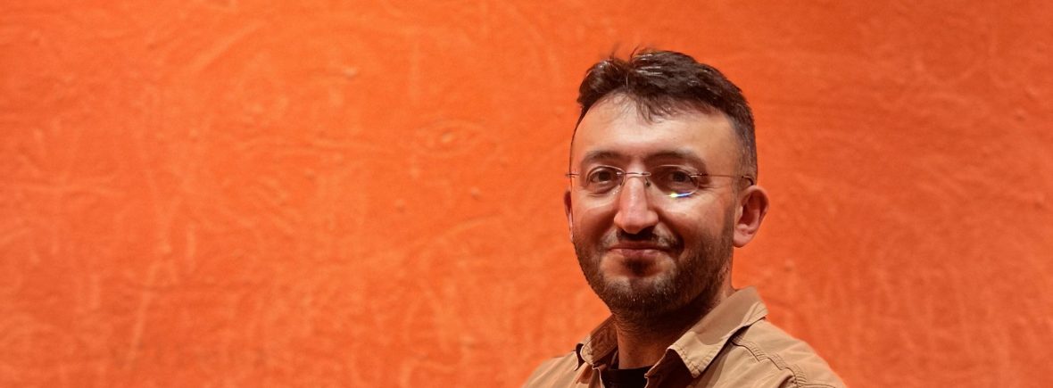 Dr. Tekin Köse - Akademik Mentor ve Ekonomist, Brighton Üniversitesi Tekin Kose wearing glasses and a brown shirt stands in front of a textured orange wall, smiling softly at the camera. Dr. Tekin Köse Akademik Mentor ve İktisatçı Türkiye